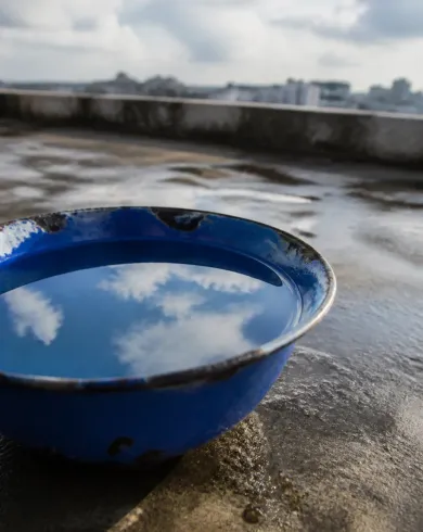 自然光類比攝影／實體佈景作品，雨後屋頂上的藍色琺瑯碗，水面映著白雲與遠方天空。直幅構圖保留清楚主體與手作質地。