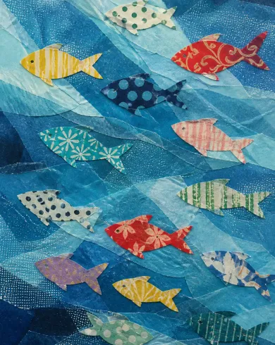 手工紙藝／立體拼貼作品，描繪不同花色包裝紙剪成魚群，游過層層半透明藍色薄紙。直式構圖保留清晰主體、自然材質與光線細節。