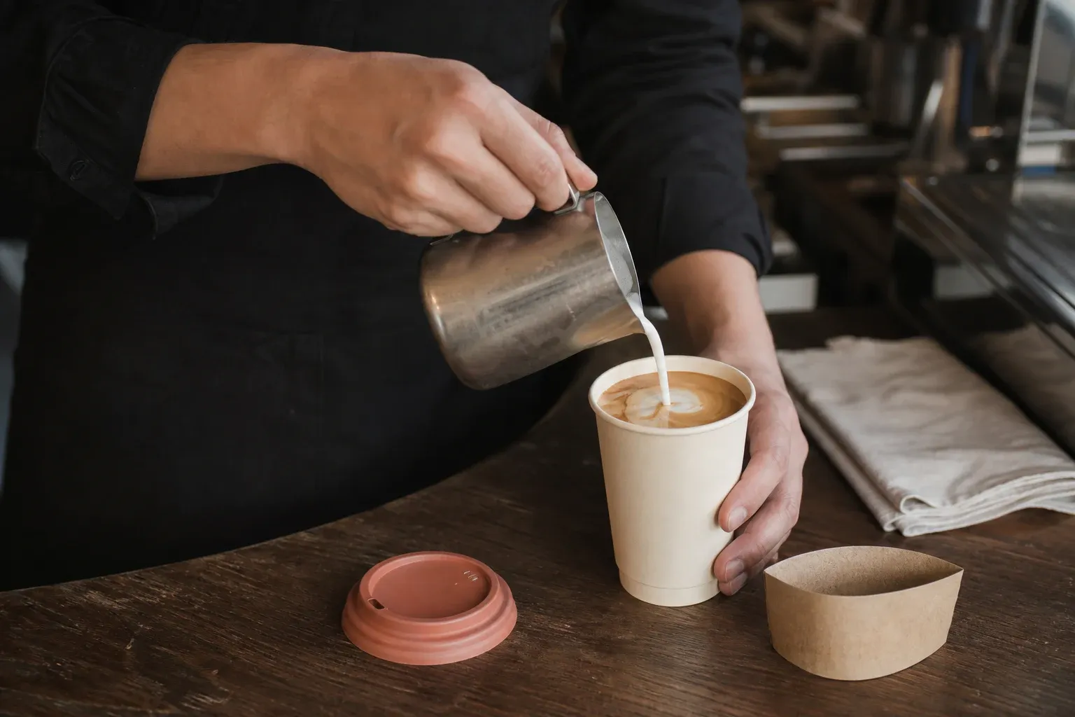 咖啡師以金屬拉花杯將鮮奶倒入紙杯拿鐵，木桌旁放著杯蓋與外帶餐盒