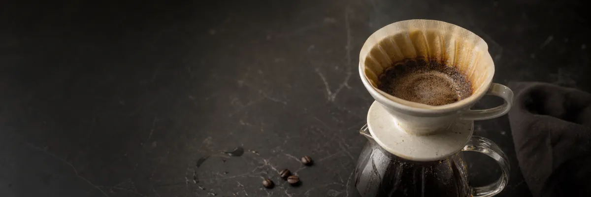 Coffee brewing through a ceramic dripper into a glass server beside scattered beans