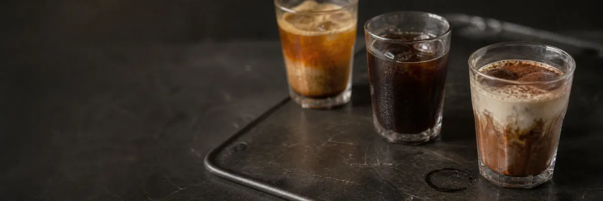 Three iced signature coffees in amber, black and cream tones on a dark metal tray